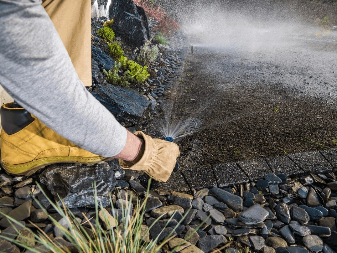A person adjusting a sprinkler head while water sprays onto a landscaped area.